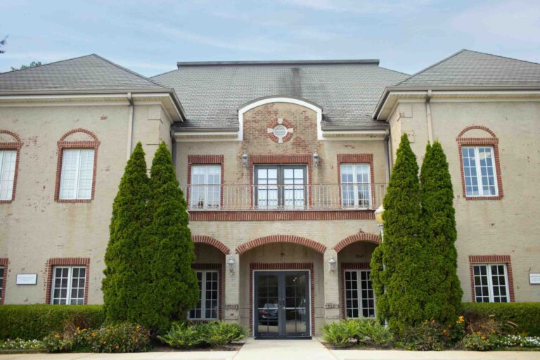 A large beige building with red brick accents and a gray roof. It features a symmetrical design, tall evergreen trees flanking double glass doors, and a balcony above the entrance with neatly trimmed bushes and flowers in the foreground.