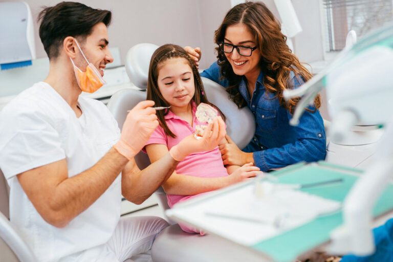 doctor showing a child patient a jaw model with the patient's mother behind them smiling