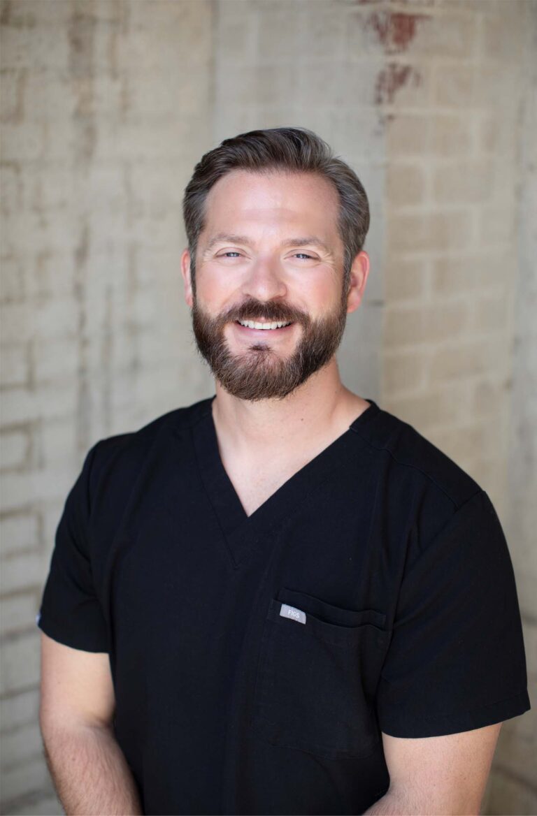 Headshot of Dr. Hall stands against a brick wall wearing a black V-neck scrub top with a small pocket on the chest.