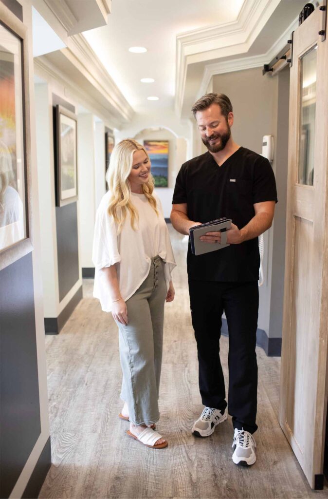 Two individuals stand in a hallway, one in white attire with sandals and Dr. Hall wearing black attire with sneakers, interacting using a tablet in a well-lit environment with framed artwork on the walls.