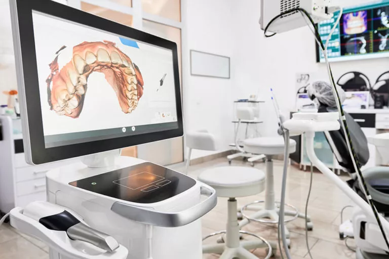 The image shows a modern dental office featuring a digital dental scanner and a computer screen displaying a 3D model of a dental arch.