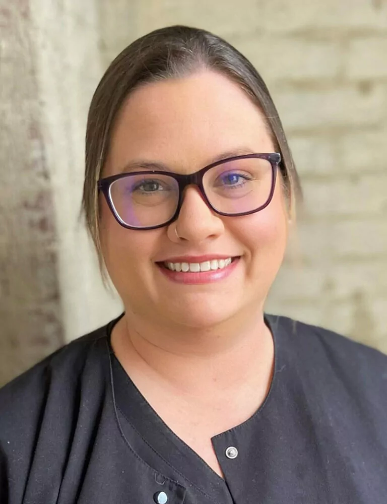 Headshot of Courtney wearing a black collared shirt stands against a light brick wall. Their hair is neatly tied back, and they wear rectangular glasses that add a thoughtful, composed touch to their appearance.