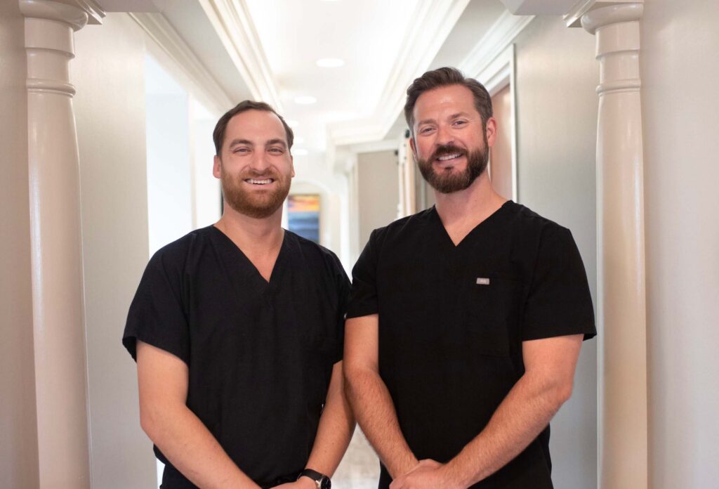 Dr. Hall and Dr. Alison, wearing black scrubs, stand side by side in a well-lit hallway, with artwork visible in the background.