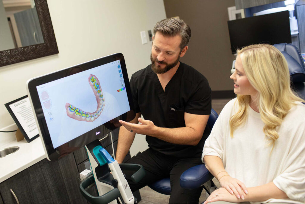 Dr. Hall in a clinic explains a 3D dental scan displayed on a computer screen to a patient seated in a dental chair. The scan showcases a colorful model of dental structures.