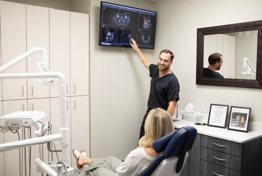 In a dental office, a patient seated in a dental chair listens as Dr. Alison points at a wall-mounted screen displaying X-rays or scans. Cabinets, equipment, and other items are visible in the room.