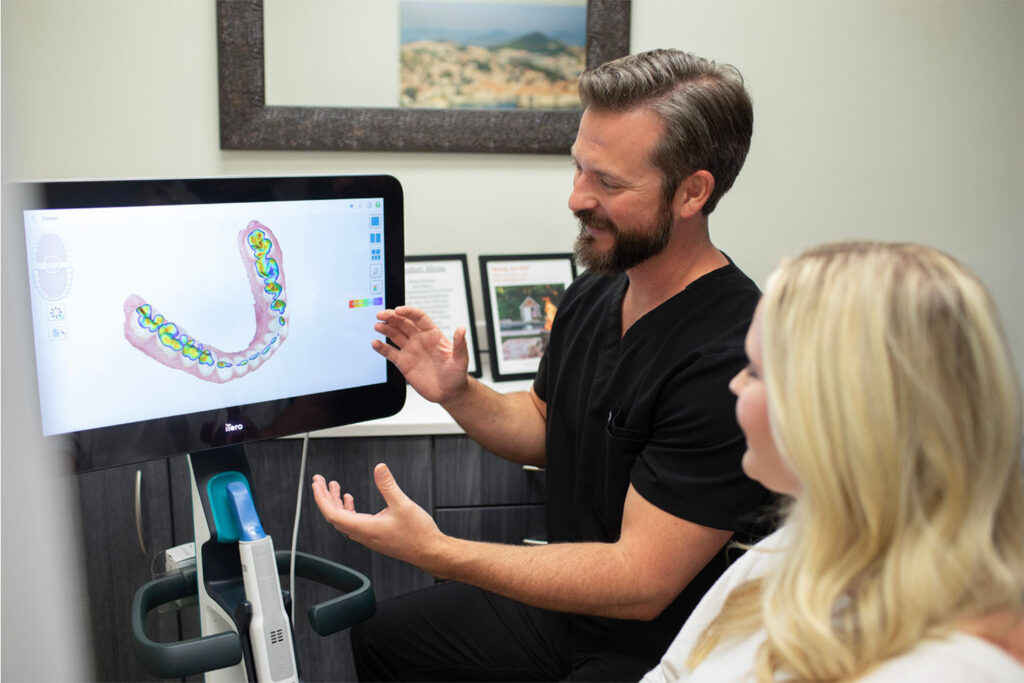 Dr. Hall points to a digital 3D model of a lower jaw displayed on a screen, highlighting areas with different colors in a clinical consultation room.
