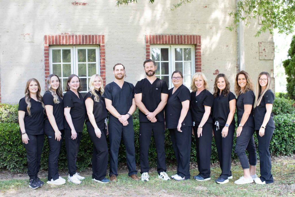 Eleven individuals dressed in black uniforms stand in front of a light-colored building with brick-bordered windows and surrounding greenery, likely a team group photo in a professional setting.